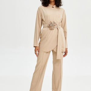 Puede incluir: Un traje de pantalón beige con una chaqueta de manga larga y pantalones de pierna ancha. La chaqueta tiene un cuello mandarín y una hebilla de cinturón decorativa con forma de flor.