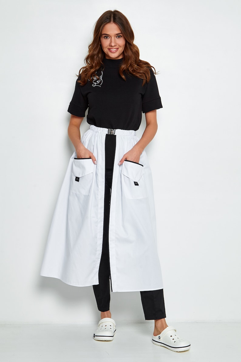 May include: A white maxi skirt with a black waistband and two large pockets on the front. The skirt has a black zipper down the front and a black belt with a silver buckle. The model is wearing a black mock neck top and black trousers.