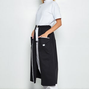 May include: A black long skirt with white piping and two large pockets on the front. The skirt has a white elastic waistband and a zip closure. The skirt is worn with a white mock neck shirt and white shoes.