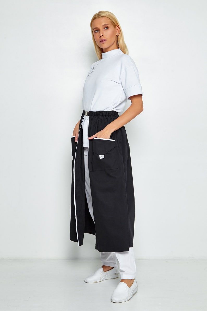 May include: A black long skirt with white piping and two large pockets on the front. The skirt has a white elastic waistband and a zip closure. The skirt is worn with a white mock neck shirt and white shoes.