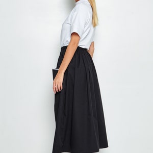 May include: A black midi skirt with a white trim and a white pocket on the side. The skirt is pleated and falls to the ankles. The model is wearing a white t-shirt and white trainers.