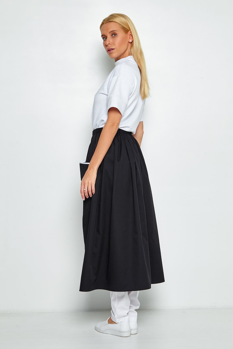 May include: A black midi skirt with a white trim and a white pocket on the side. The skirt is pleated and falls to the ankles. The model is wearing a white t-shirt and white trainers.