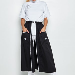 May include: A black skirt with white piping and two pockets. The skirt is worn over white trousers and a white mock neck shirt with a line drawing of a face on the front.