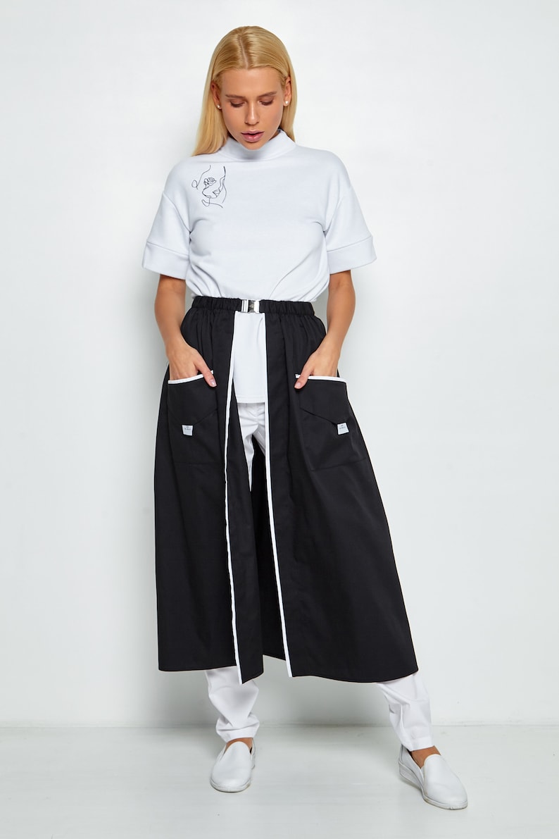 May include: A black skirt with white piping and two pockets. The skirt is worn over white trousers and a white mock neck shirt with a line drawing of a face on the front.