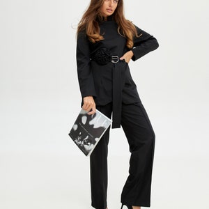 Puede incluir: Una camisa negra de cuello alto con un detalle de flor de tela negra en la parte delantera. La camisa tiene un cinturón negro con una hebilla plateada. La modelo lleva pantalones negros de pierna ancha y tacones negros. La modelo sostiene una revista que dice "Vogue" en la portada.