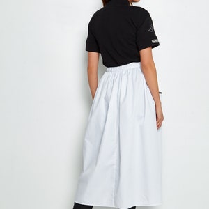 May include: A white skirt with a gathered waistband and a black mock neck shirt. The shirt has a small graphic on the sleeve. The person is wearing black trousers and white shoes.