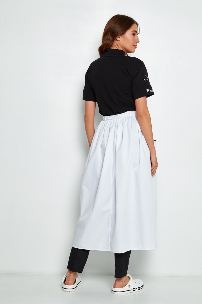 May include: A white skirt with a gathered waistband and a black mock neck shirt. The shirt has a small graphic on the sleeve. The person is wearing black trousers and white shoes.