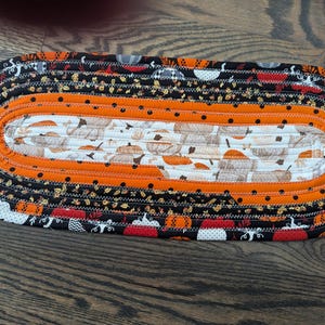 Könnte beinhalten: Ovaler, gesteppter Tischläufer mit Herbstthema. Der Läufer hat ein zentrales Feld mit Kürbismustern, umgeben von orangefarbenen, schwarzen und gemusterten Stoffrändern. Die Kanten sind mit einem Zickzackstich versehen, was einen dekorativen Akzent schafft.