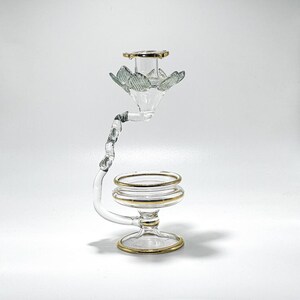 May include: A clear glass candle holder with gold edging. The candle holder features a distinctive, curved design, with a flower-shaped top and a circular base. The gold edging adds a touch of elegance to the piece.