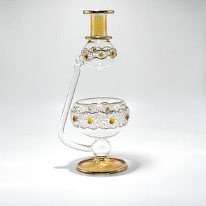 May include: A clear glass decorative piece with gold accents. The piece has a bulbous base and a top section, connected by a curved glass tube. Gold trim and amber-coloured embellishments adorn the glass, creating an elegant design.