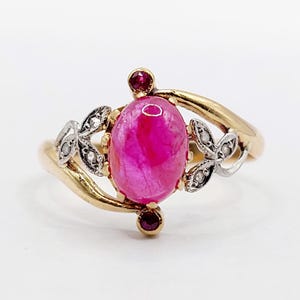 May include: A gold ring featuring a large, oval, pink gemstone. The ring has two smaller red gemstones and two silver-colored, leaf-shaped accents with small, clear stones. The band is gold.