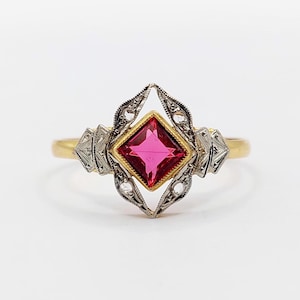 Op de afbeelding: Een gouden ring met een vierkant geslepen, felroze edelsteen in een geelgouden zetting. De middelste steen wordt omlijst door een ingewikkelde, puntige zilverkleurige metalen zetting met decoratieve gravures en uitgesneden details.