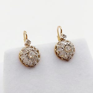 May include: A pair of gold drop earrings featuring spherical, openwork cages with a star-like pattern. Each earring is suspended from a small, square-shaped setting containing a single clear stone, attached to a curved wire hook closure.