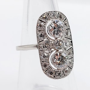 May include: A silver ring with an oval face, featuring two large, round diamonds and numerous smaller diamonds. The ring's design is symmetrical, with the diamonds set in a geometric pattern. The ring is displayed against a clear background.