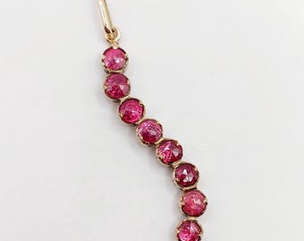 May include: A gold pendant featuring a curved vertical row of seven round, faceted, deep pinkish-red stones set in individual metal prongs. The piece includes a small gold bail at the top for attachment to a chain or necklace.