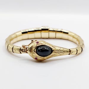 Puede incluir: Una pulsera dorada con forma de serpiente y banda segmentada flexible. La cabeza de la serpiente presenta patrones grabados, dos pequeños ojos de piedra roja y una gran piedra negra ovalada de tipo cabujón engastada en el centro.