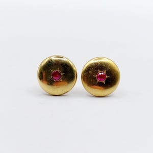 Op de afbeelding: Een paar goudkleurige oorstekers. Elke oorsteker heeft een ronde, gouden basis met een kleine, rode edelsteen in het midden. De edelstenen zijn omgeven door een sterrenpatroon. De oorbellen staan tegen een witte achtergrond.