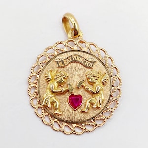 Peut inclure: Pendentif en or avec une bordure en filigrane. Le pendentif représente deux chérubins encadrant un rubis en forme de cœur. Le texte "IL BAT POUR TOI" est inscrit sur une bannière au-dessus des chérubins.