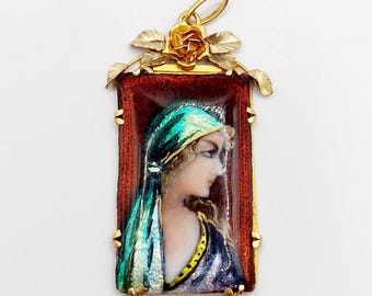 Puede incluir: Colgante dorado con un retrato de una mujer de perfil, con un pañuelo turquesa y una prenda azul oscuro y dorada. El retrato está enmarcado en un marco rectangular con fondo rojo y borde dorado. Una rosa y hojas doradas adornan la parte superior.