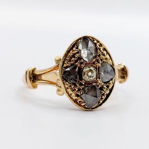 Op de afbeelding: Een gouden ring met een ovaalvormig blad met een textuur van kruisarceringen. Het midden is bezet met een kleine, lichtgekleurde steen, omringd door vier grotere, donkere, gefacetteerde stenen in een kruispatroon op de gouden ring.