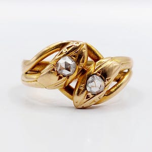 May include: A gold ring featuring two intertwined snake heads, each set with a single clear rose-cut diamond. The band is crafted from polished yellow gold, with the snake heads meeting in the centre, showcasing detailed scales and eyes.