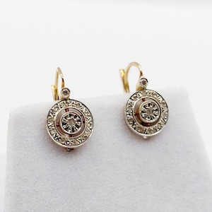 May include: A pair of gold leverback earrings featuring a circular design with a central round stone surrounded by an intricate, engraved metal border. The earrings have a polished finish and a delicate, openwork structure that highlights the craftsmanship.