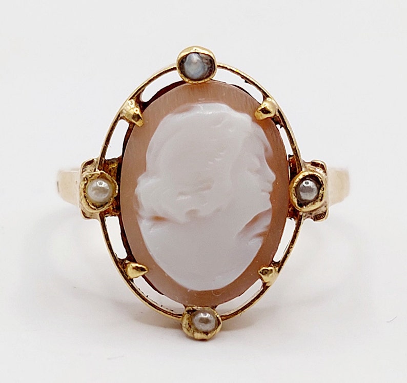 May include: Gold ring with a cameo featuring a woman's profile. The cameo is set in a gold frame with three small pearls on each side.