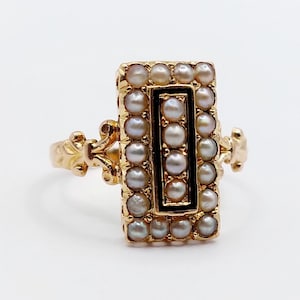 Puede incluir: Un anillo de oro con una cara rectangular engastada con pequeñas perlas blancas que rodean una incrustación central de esmalte negro. El engaste rectangular está bordeado por una hilera de perlas y el aro presenta detalles metálicos.