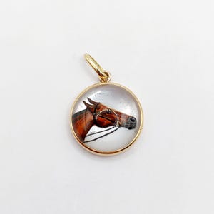 May include: A gold-toned pendant featuring a round glass cabochon with a painted image of a brown horse's head. The horse is wearing a bridle and is looking to the right.