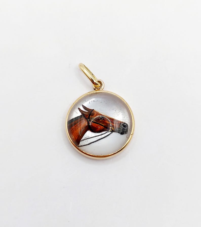 May include: A gold-toned pendant featuring a round glass cabochon with a painted image of a brown horse's head. The horse is wearing a bridle and is looking to the right.