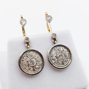 May include: A pair of drop earrings featuring a circular pendant design with a central round diamond surrounded by smaller diamonds in a cluster setting. Each earring has a vertical bar with two additional small diamonds and a lever back closure.