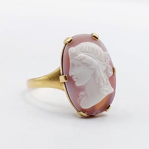 May include: A gold ring featuring an oval-shaped cameo with a white profile portrait of a person carved against a soft pink background. The cameo is secured by six small gold prongs, and the band is a smooth, polished gold metal finish.