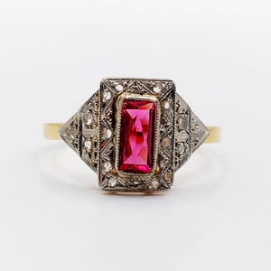 May include: A gold ring featuring a rectangular, vibrant pink gemstone set in a silver-toned, filigree-style mounting. The setting is decorated with small, clear, round stones and intricate metalwork, extending into a tapered, pointed design.