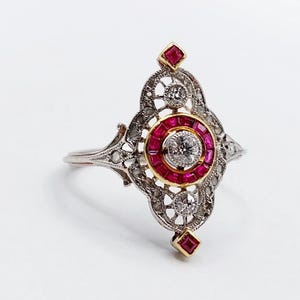 May include: An antique ring with a silver band and a detailed filigree design. The ring features a central round diamond surrounded by a halo of small rubies. Additional diamonds and rubies are set in the ornate setting.