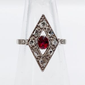 May include: A silver-toned ring featuring a central oval-cut red gemstone set within a diamond-shaped frame. The frame is embellished with multiple small, clear, faceted stones arranged in a geometric pattern, with a thin, polished metal band.