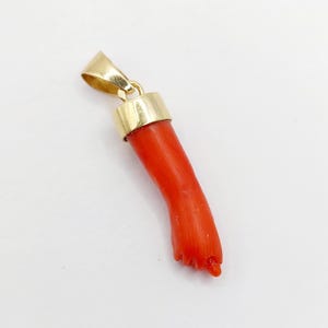 May include: A pendant featuring a carved red coral figa hand charm suspended from a polished yellow gold bail. The coral is smooth and vibrant, shaped into a clenched fist with the thumb tucked between the index and middle fingers, set in gold.