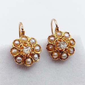 May include: A pair of gold earrings featuring a central clear faceted stone surrounded by a circular cluster of small, round, iridescent white pearls. Each earring is designed with a curved wire hook for attachment, showcasing a floral shape.