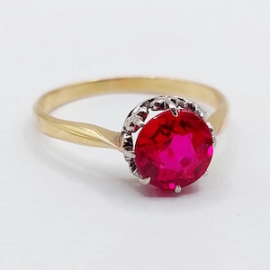 May include: A gold ring with a large, round, bright red gemstone. The gemstone is set in a silver-colored bezel, and the band is a simple, polished gold. The ring is set against a white background.