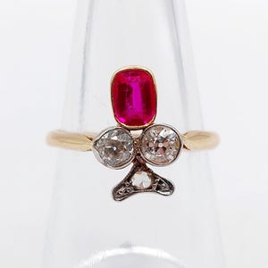 May include: A gold ring featuring a central rectangular red gemstone set vertically above two round white diamonds, with a smaller triangular diamond accent at the base, all arranged in a clover-like shape on a polished gold band.