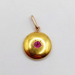 May include: A gold pendant with a round, polished surface. The pendant features a central, bright pink gemstone set within a starburst design. The pendant has a small loop for attaching to a chain.