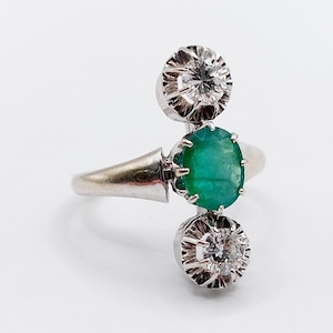 May include: A silver ring featuring a central oval emerald gemstone, flanked by two round, sparkling diamonds. The ring's design is elegant, with a unique arrangement of the gemstones. The ring is set against a plain white background.