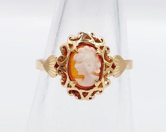 Op de afbeelding: Goudkleurige ring met een ovale camee met een profiel van een vrouw. De camee is gezet in een sierlijk, filigraan-stijl frame. De ring heeft een delicate band met decoratieve accenten aan weerszijden van de camee. Afmetingen in centimeters.