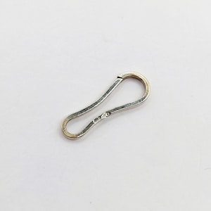 Puede incluir: Un cierre plateado y alargado con un diseño curvo. El cierre tiene forma de gancho en un extremo y un lazo redondeado en el otro. El metal tiene un aspecto ligeramente texturizado, sobre un fondo blanco.