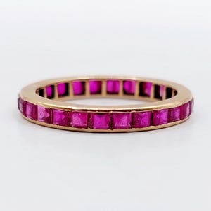 Puede incluir: Un anillo de oro con una banda de piedras preciosas rosas vibrantes, talladas en forma cuadrada. Las piedras están engastadas muy juntas, creando una línea continua de color alrededor del anillo. Sobre un fondo blanco.