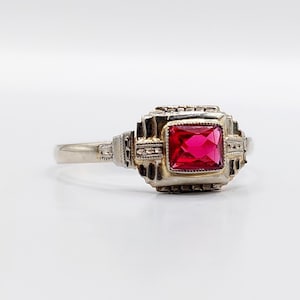 Peut inclure: Une bague en argent ornée d'une pierre rouge vif de forme carrée. Le sertissage de la bague présente un design Art Déco avec des détails complexes. La monture est fine et simple, contrastant avec la pièce maîtresse ornée. La bague est présentée sur une surface blanche.
