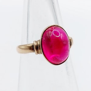May include: A gold ring featuring a large, oval-shaped, vibrant pink gemstone. The ring has a simple band with decorative detailing near the gemstone setting. The gemstone is the focal point, showcasing a polished, smooth surface.