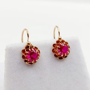 May include: A pair of gold drop earrings featuring a central round-cut vibrant pink gemstone. Each stone is held in a prong setting surrounded by a scalloped, petal-like gold frame. The earrings are attached to curved gold ear wires for hanging.
