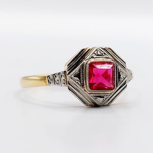 Peut inclure: Une bague vintage avec une pierre rouge vif carrée, sertie dans un cadre géométrique argenté. La monture est dorée et la bague présente des détails complexes sur les épaules.