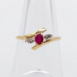 Pode incluir: Um anel de ouro com um rubi vermelho central de corte oval numa cravação de quatro garras. A banda tem um design cruzado com dois pequenos detalhes prateados em forma de folha de cada lado do rubi, cada um com uma pequena pedra.