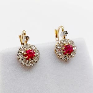 Puede incluir: Un par de pendientes de oro con un rubí rojo central de talla redonda rodeado por un halo de pequeñas piedras transparentes. Los pendientes tienen un cierre de palanca y están engastados en una combinación de tonos de metal amarillo y blanco.